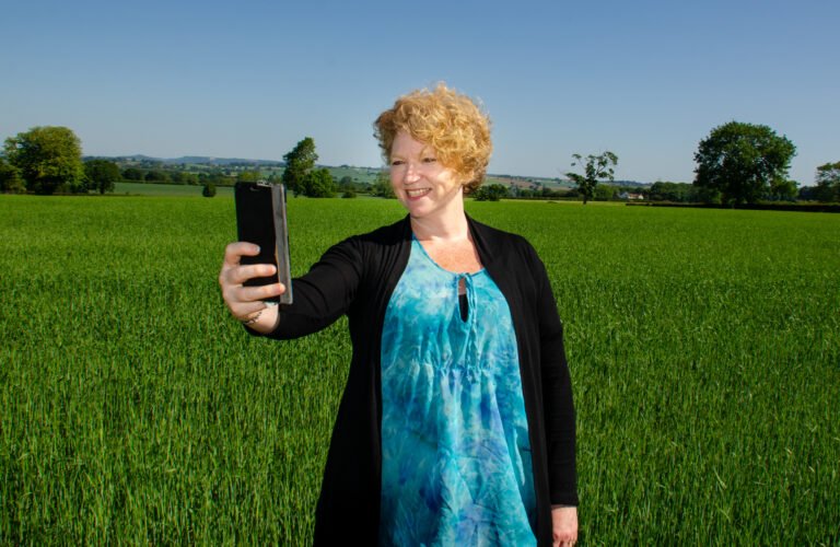Jessa singing to her phone, in a green field. Jessa is dressed in a turquoise floaty top and black cardigan.