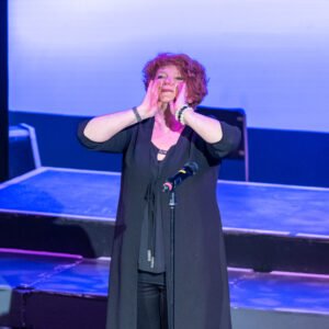 Jessa with her hands at the sides of her mouth, calling out to a theatre audience.